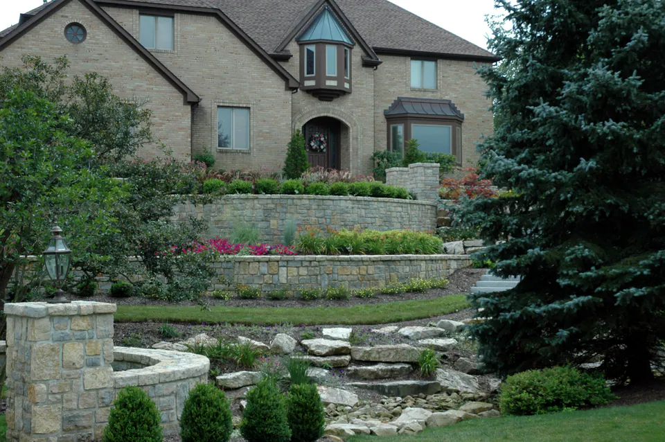 Curved stone retaining walls with pink annuals, boulders, and evergreens in front of a brick house